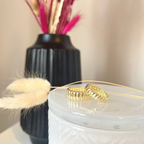 Gold rings on a white textured surface with a black vase and pink feathers in the background.