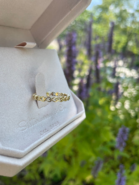 A delicate gold ring with a wovenh heart pattern, resting on a white jewelry box cushion. The background is a soft-focus outdoor garden with green foliage and purple flowers.