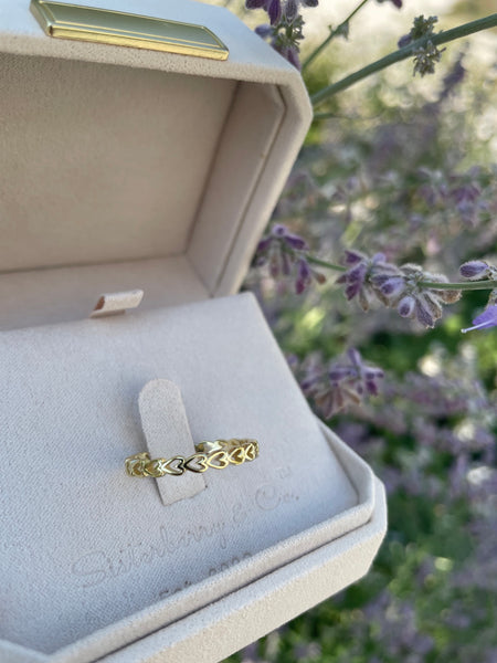 A gold ring with an intricate heart pattern, displayed on a light beige velvet ring box. The box is open, and the background is a soft-focus outdoor garden with purple flowers.