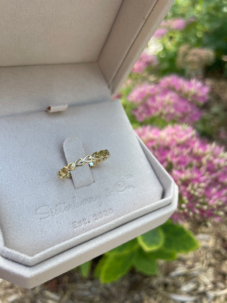 A delicate yellow gold ring with a heart pattern, displayed on a gray beige ring box with the brand name 'Sisterberry & Co.' with pink floral garden in the backround