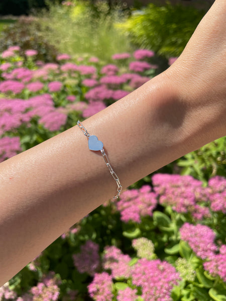 A delicate silver chain bracelet with a small heart charm, worn on a person's wrist against a blurred background of pink flowers and green foliage.