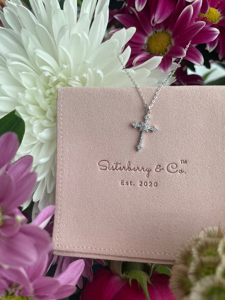 A silver cross pendant with pave cubic zirconia, displayed on a pink jewelry pouch embossed with Sisterberry & Co. logo.