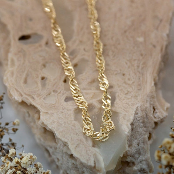 Gold chain bracelet on a textured beige surface with dried flowers.