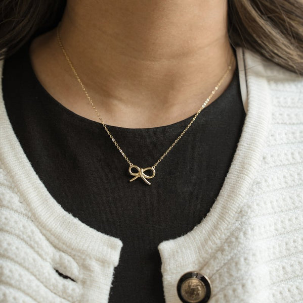 Gold bow necklace worn by a person with a black shirt and white cardigan.