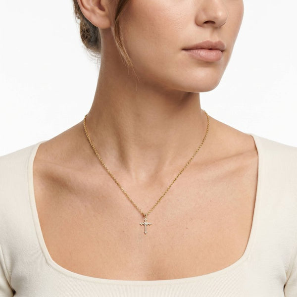 A close-up of a model wearing a delicate gold chain necklace with a small cross pendant, styled with a cream-colored scoop-neck top against a clean white background.