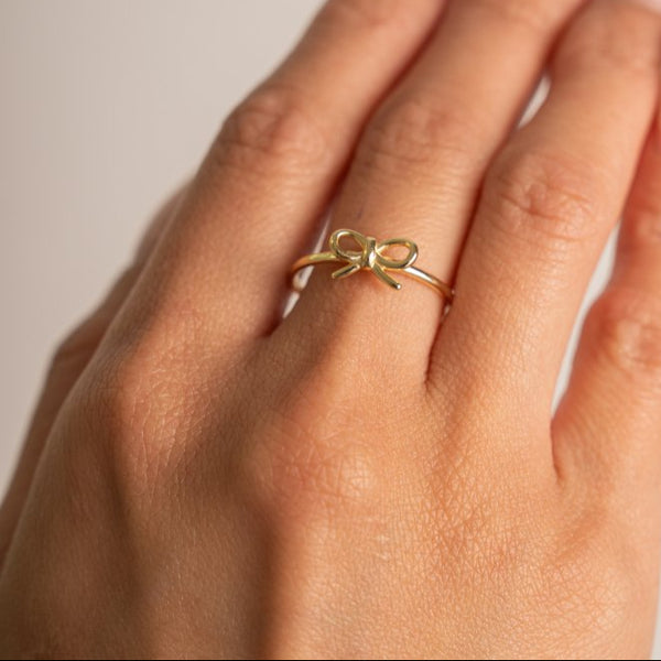 Gold bow-shaped ring on a hand against a neutral background