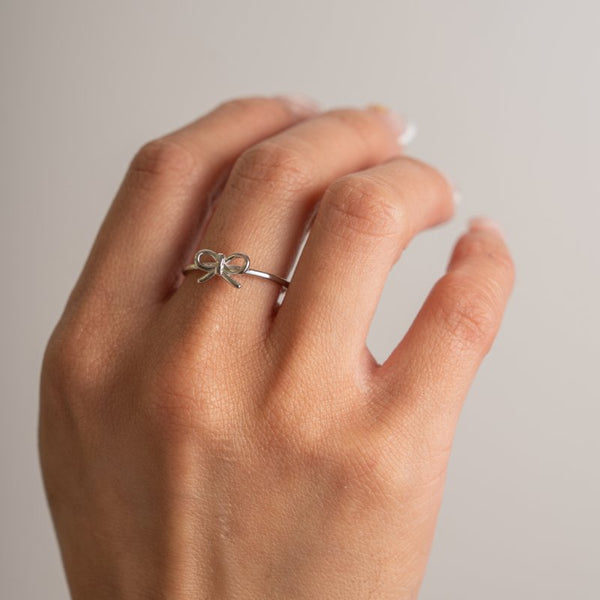 Silver bow-shaped ring on a person's finger against a neutral background